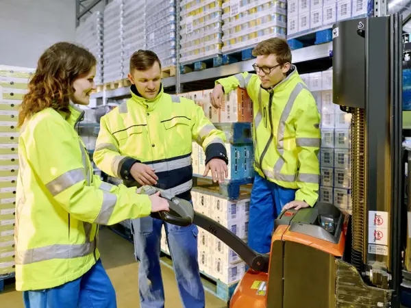 Zwei Auszubildende der Logistik stehen mit ihrem Ausbilder im Kühllager an einer Logistik-Ameise und lernen.