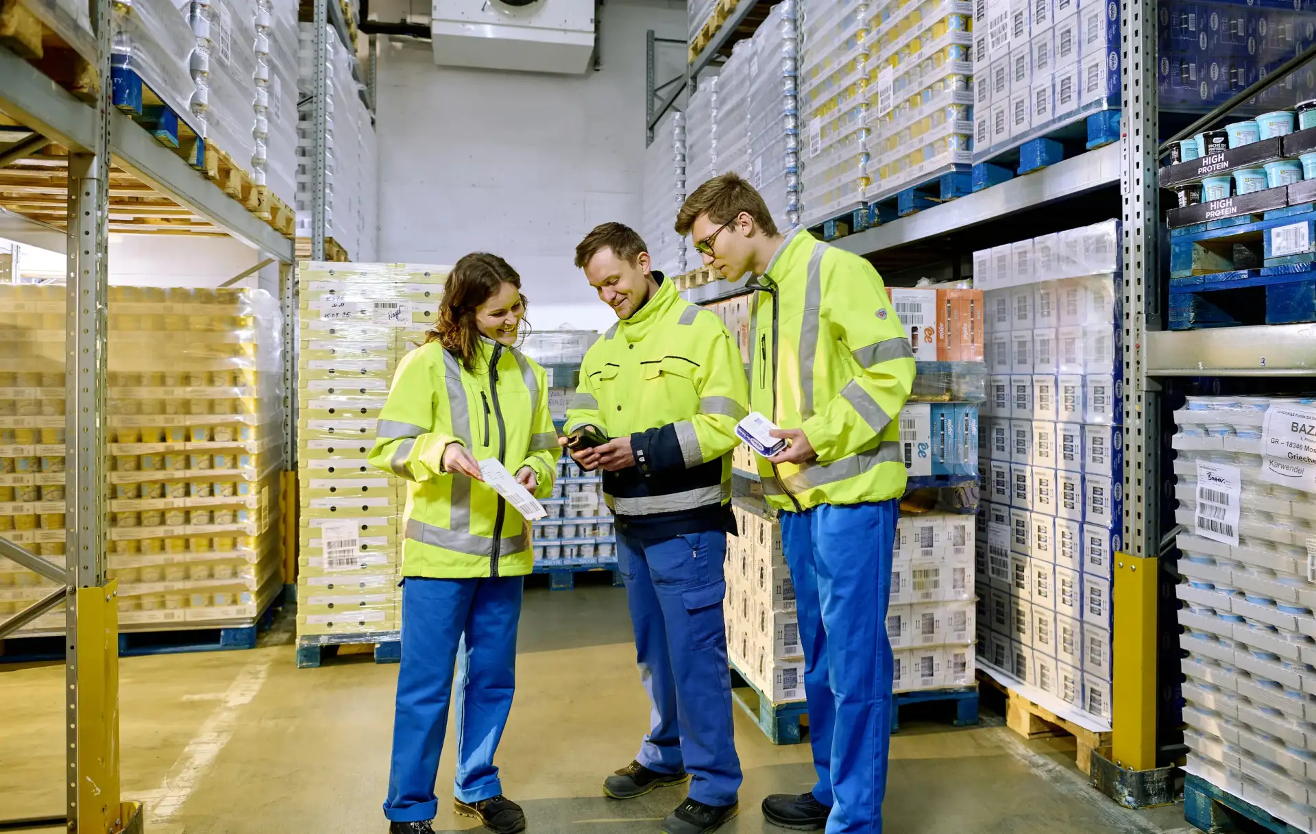 Zwei Auszubildende der Logistik stehen mit ihrem Ausbilder im Kühllager und erhalten eine Erklärung.