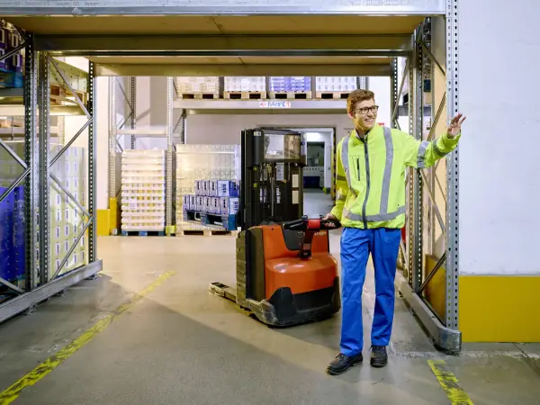 Ein Logistik-Azubi zieht eine Ameise durchs Lager und winkt Kollegen zu.