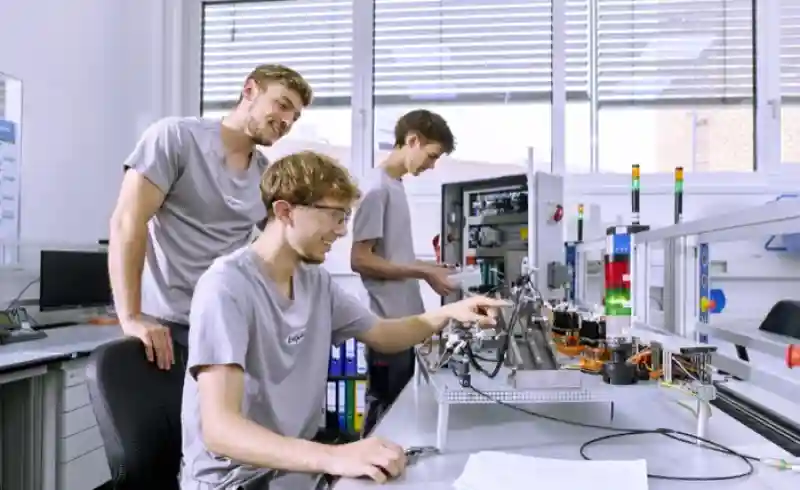 Zwei Mechatroniker-Azubis lernen am Elektropult von ihrem Ausbilder.