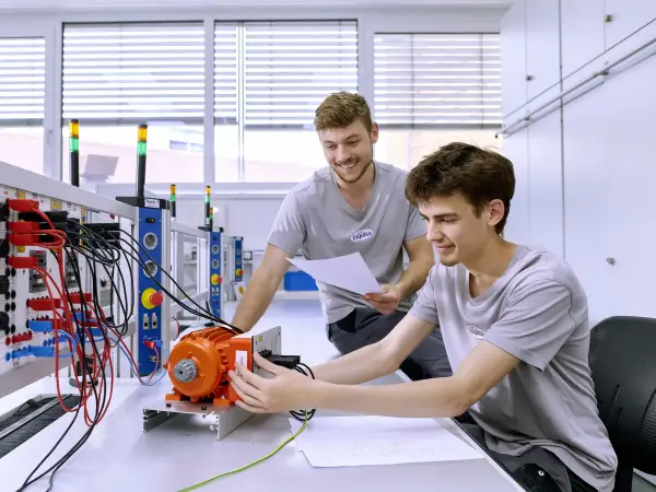 Ein Mechatroniker-Azubi arbeitet am Elektropult, während sein Ausbilder die Arbeit prüft.