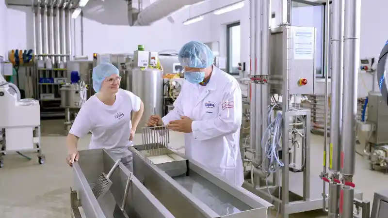 Eine Auszubildende der Produktion steht mit ihrem Ausbilder an einer Käsetheke und lernt das Käsen.
