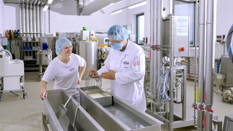 Eine Auszubildende der Produktion steht mit ihrem Ausbilder an einer Käsetheke und lernt das Käsen.