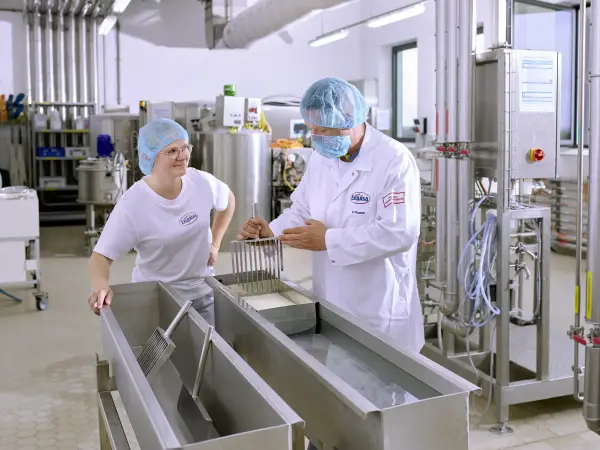 Eine Auszubildende der Produktion steht mit ihrem Ausbilder an einer Käsetheke und lernt das Käsen.