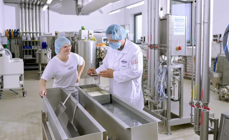 Eine Auszubildende der Produktion steht mit ihrem Ausbilder an einer Käsetheke und lernt das Käsen.