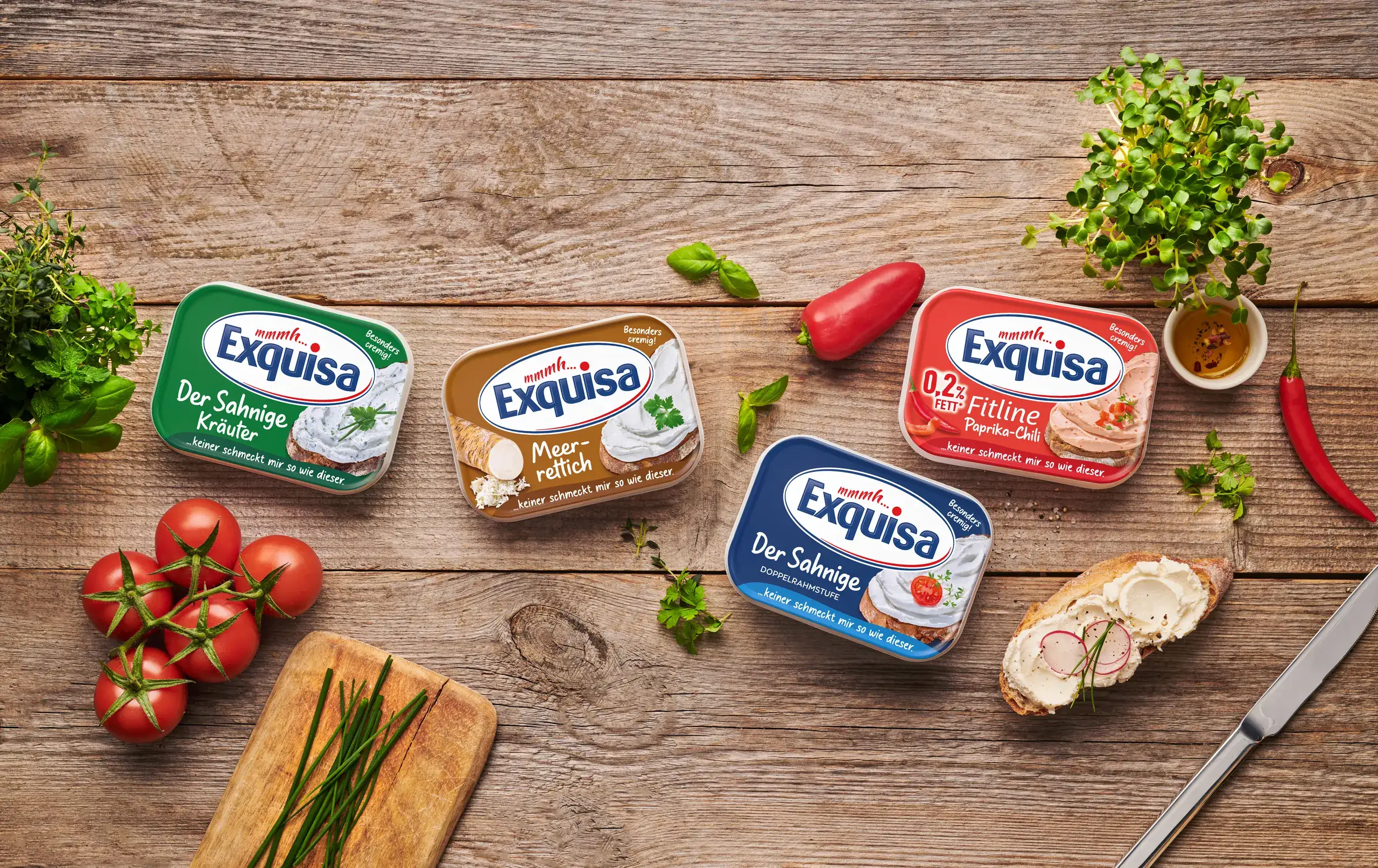 Exquisa Frischkäsesorten stehen mit Tomaten, Kräutern und Broten auf einem Holztisch.