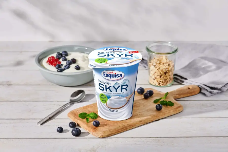 Becher eines Exquisa Milder Skyr Produkts mit Beeren und Frühstückszutaten auf einem Tisch.
