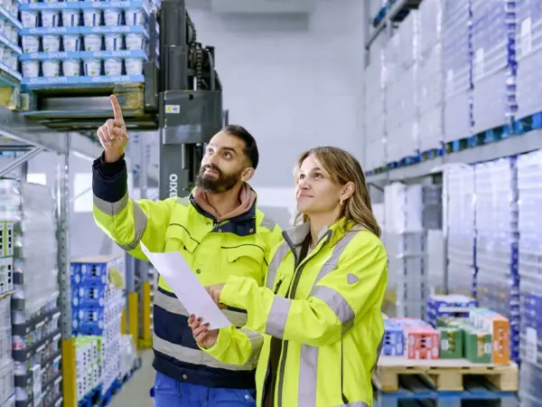 Zwei Logistikmitarbeiter in Warnwesten blicken im Kühllager nach oben vor verpackten Paletten.