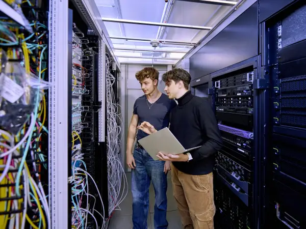 Ein IT-Mitarbeiter erklärt seinem Auszubildenden etwas im Serverraum.