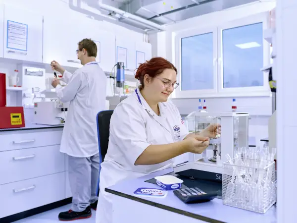 Eine Laborantin kontrolliert am Tisch die Produktqualität vor einem Kollegen im Hintergrund.