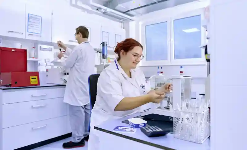 Eine Laborantin kontrolliert am Tisch die Produktqualität vor einem Kollegen im Hintergrund.