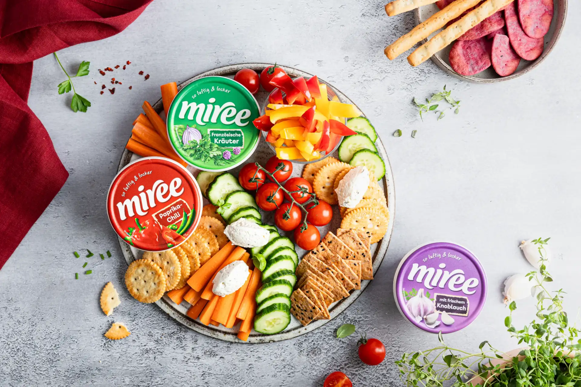 Eine gefüllte Schale steht auf einer grauen Küchenzeile. In der Schale befinden sich zwei miree Becher sowie eine Varianz an Gemüse und Dip-Snackereien wie Cracker.