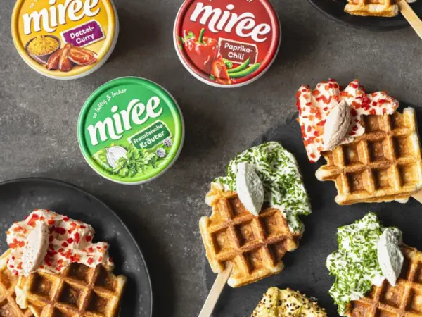 Drei miree Becher stehen mit mehreren Waffeln am Stiel auf einem grauen Tisch. Die Waffeln sind gedippt in miree und mit Kräutern, Streuseln und ähnlichem dekoriert.