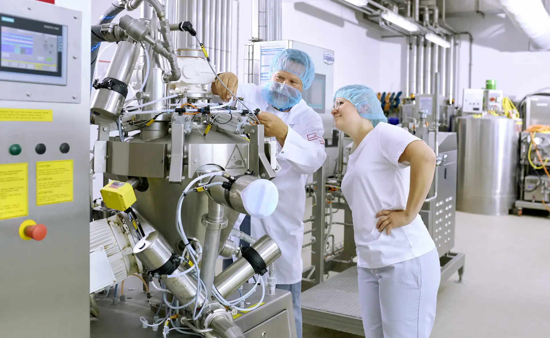 Eine Auszubildende der Produktion steht mit ihrem Ausbilder an einer Produktionsmaschine und lernt deren Bedienung.