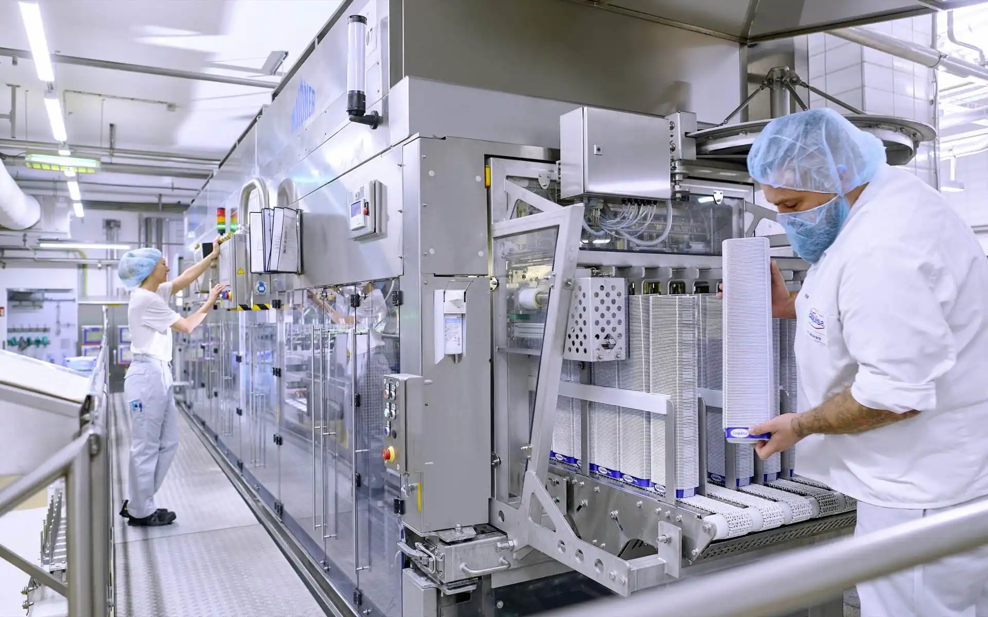 Zwei Produktionsmitarbeitende stehen an einer Produktionsmaschine. Einer bedient die Maschine und einer füllt Becher nach.