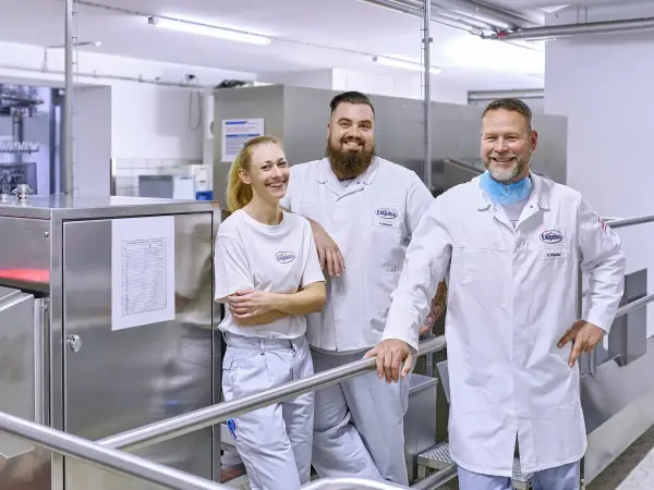 Drei Produktionsmitarbeitende jeden Alters männlich und weiblich stehen lächelnd am Eingang der Produktion.