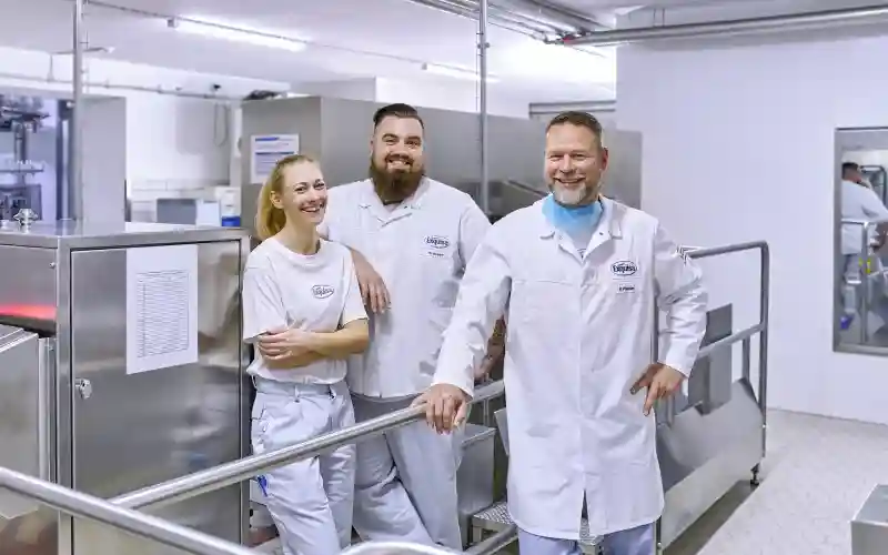 Drei Produktionsmitarbeitende jeden Alters männlich und weiblich stehen lächelnd am Eingang der Produktion.