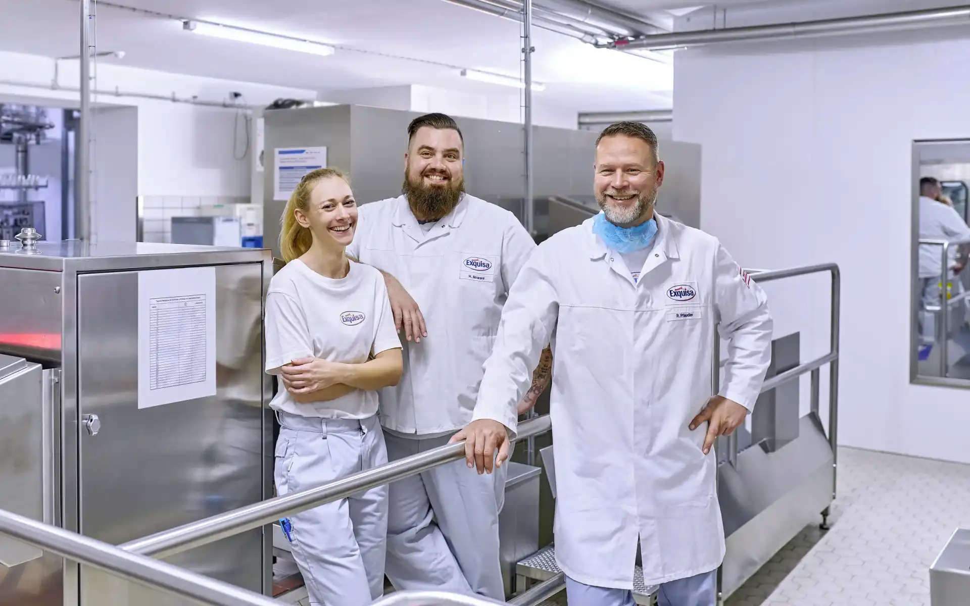 Drei Produktionsmitarbeitende jeden Alters männlich und weiblich stehen lächelnd am Eingang der Produktion.