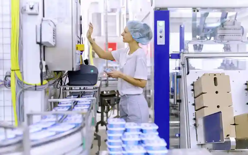 Eine Produktionsmitarbeitende steht in der Produktion an einem Laufband, auf dem Exquisa Frischkäseprodukte fahren.