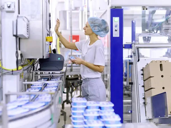 Eine Produktionsmitarbeitende steht in der Produktion an einem Laufband, auf dem Exquisa Frischkäseprodukte fahren.
